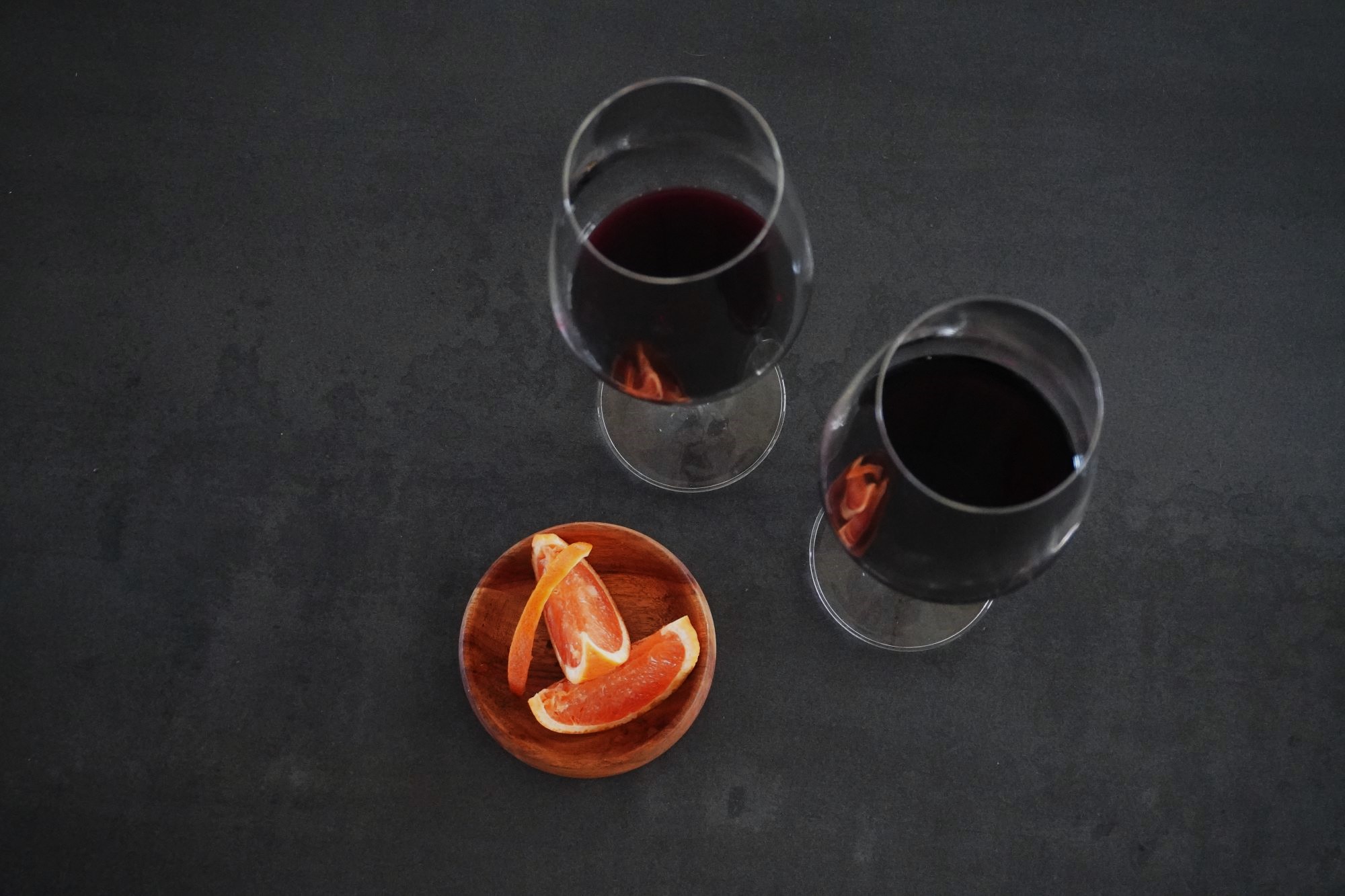 two glasses of red wine and a wooden bowl of citrus slices on a dark grey marble tabletop